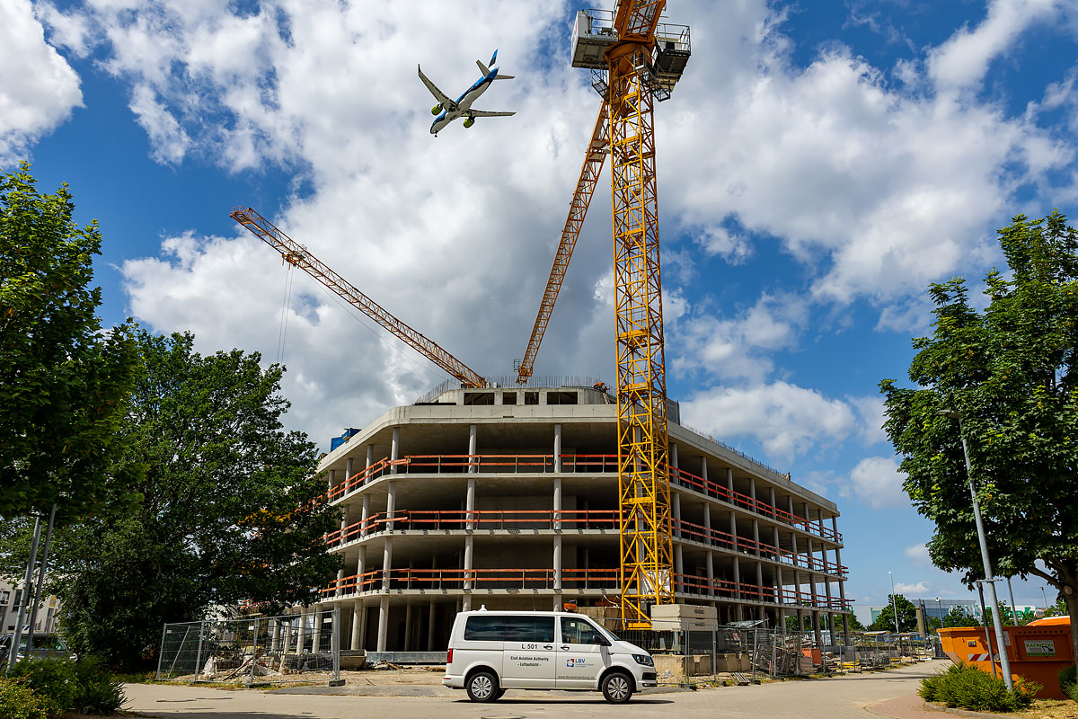 Überprüfung der Hindernisfreiheit vor einem vierstockigen Gebäude im Rohbau und einem Baukran mit einem darüberfliegenden blauweißen Flugzeug