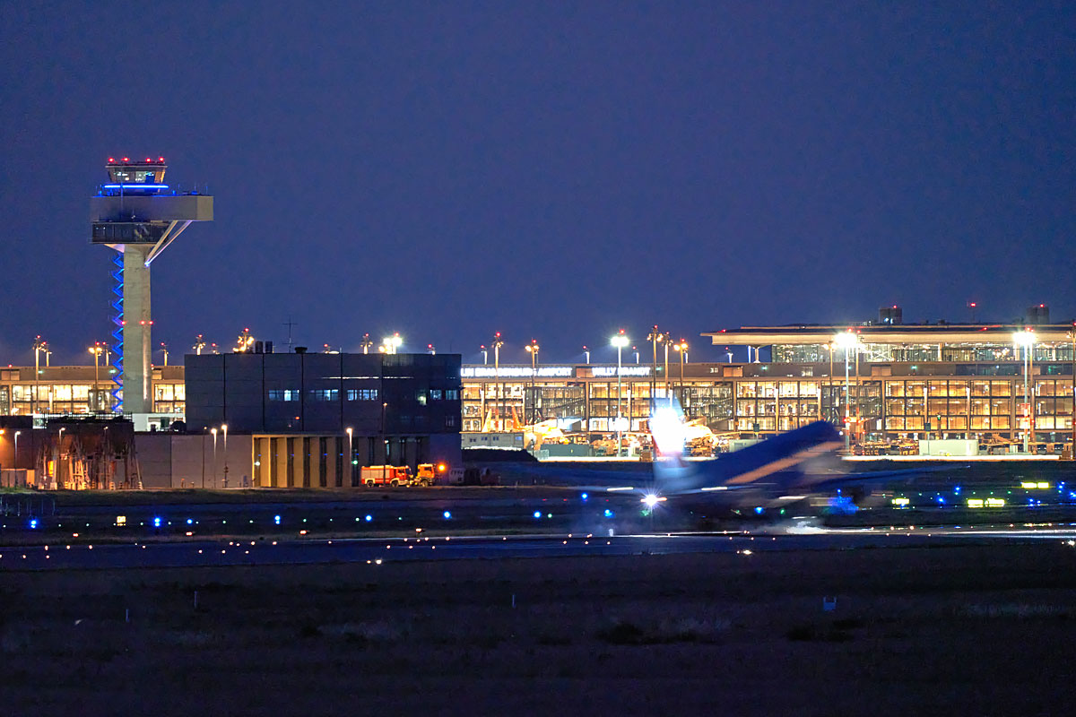 beleuchtetes Flughafengebäude des Flughafens Berlin Brandenburg bei Nacht mit einem startenden Flugzeug im Vordergrund