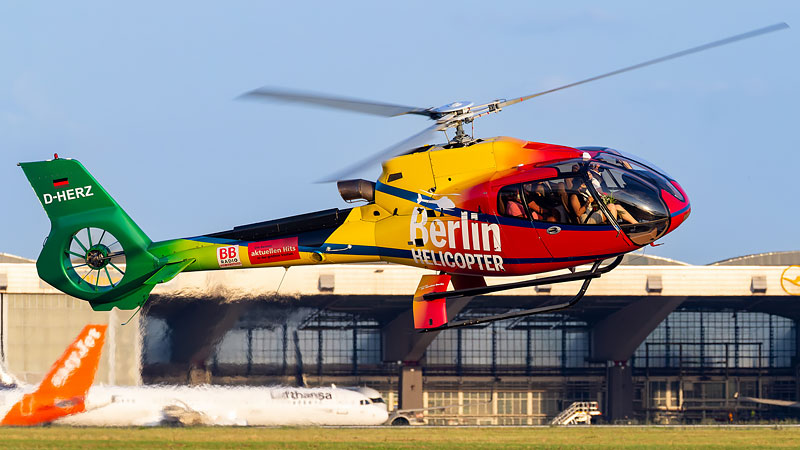 ein gelb, rot und grüner startender Helikopter im Hintergrund ein blauer Himmel und ein Flugplatzgebäude
